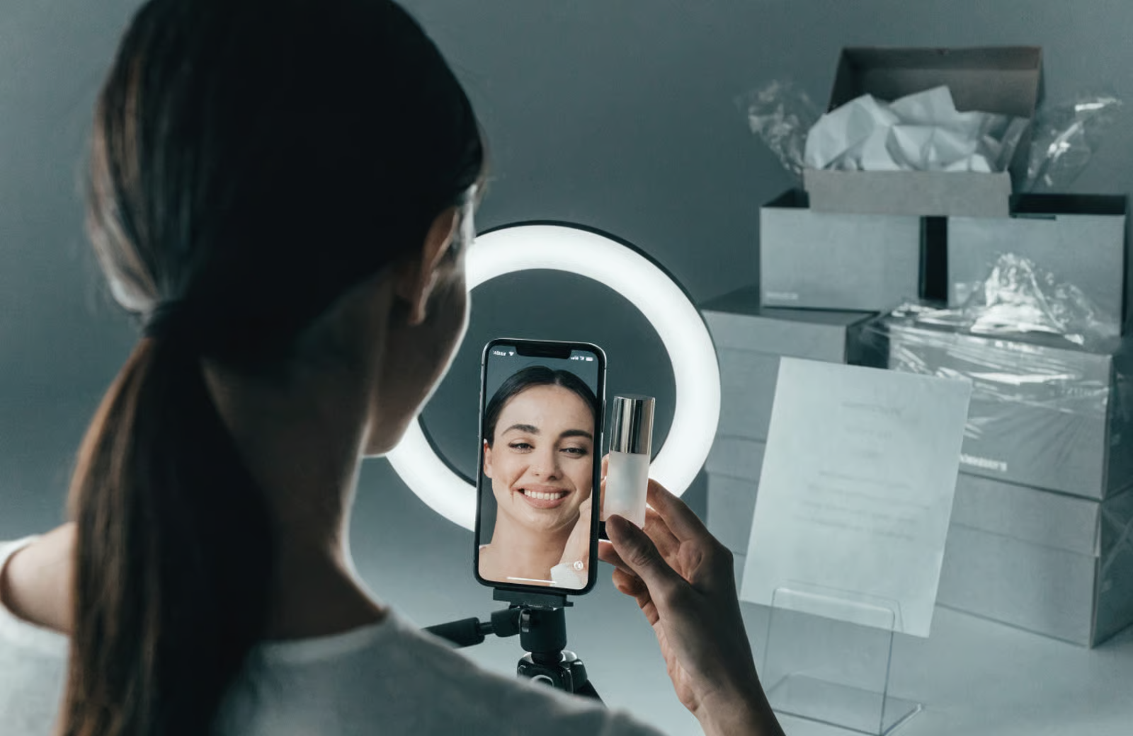 A beauty creator filming herself with a ring light and phone, holding a product, with PR boxes in the background