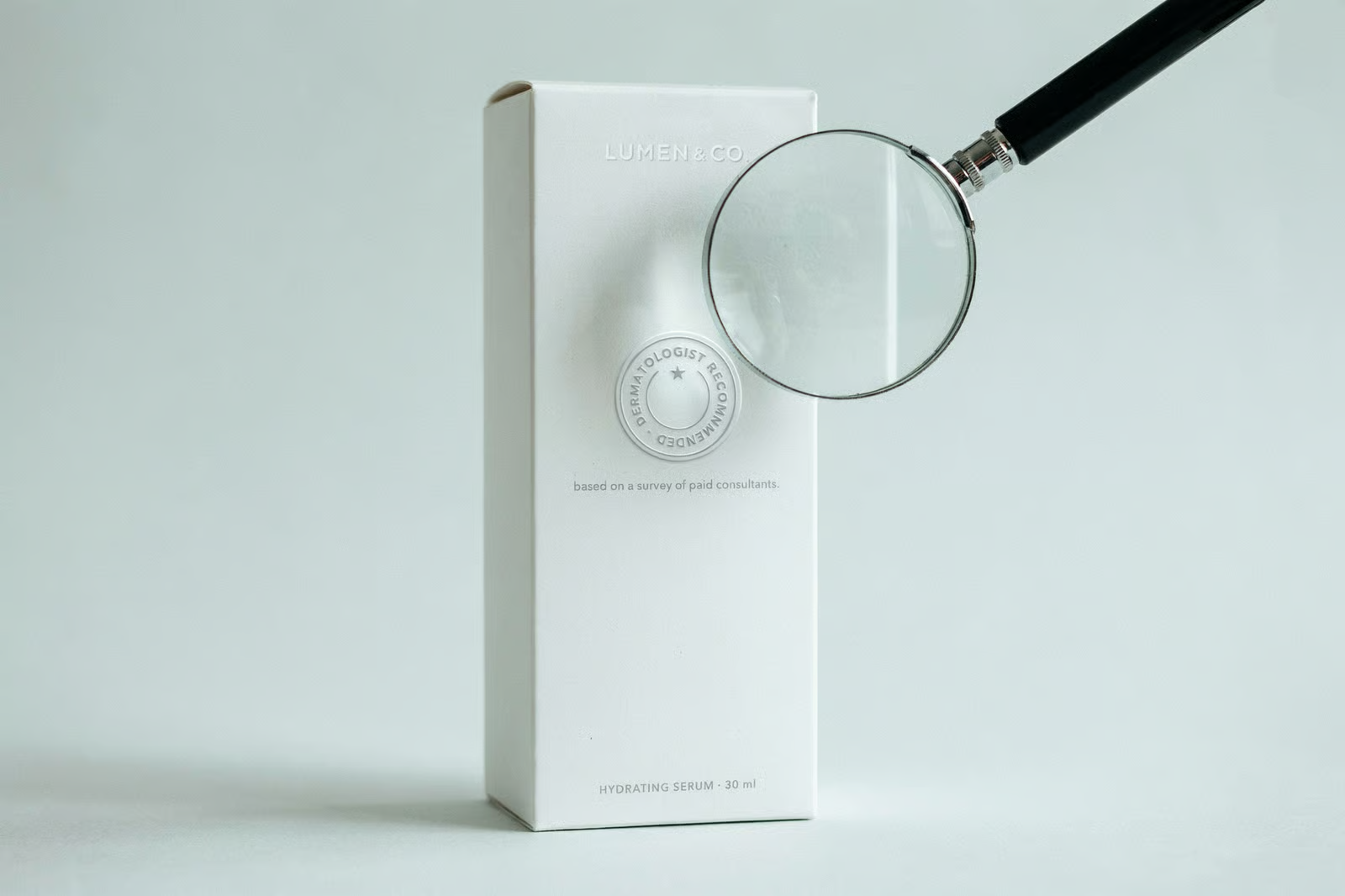 A magnifying glass held over a plain white skincare box with a small medical-style seal