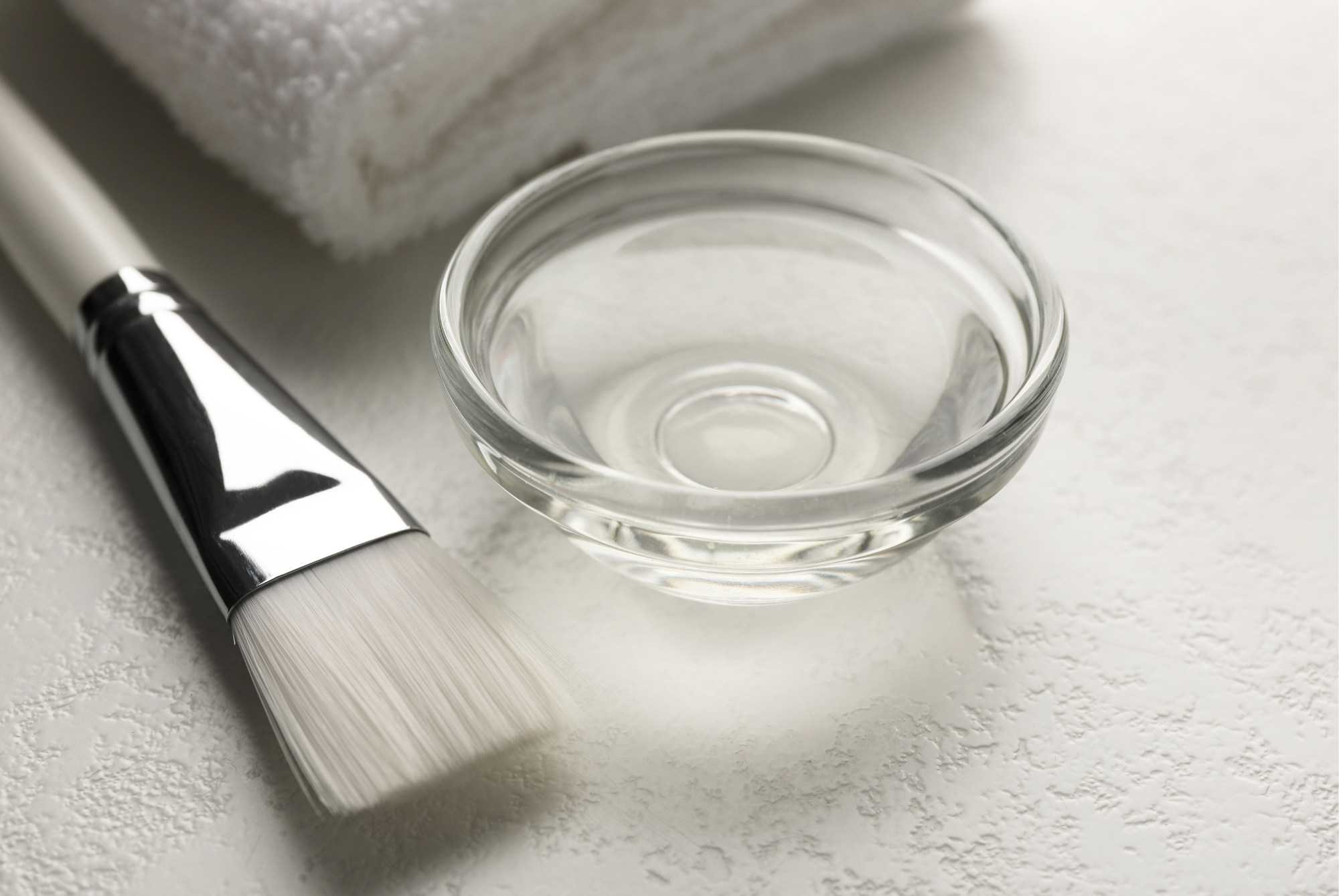 A flat-bristled applicator brush beside a small glass bowl of clear liquid on a white surface, with a folded white towel behind