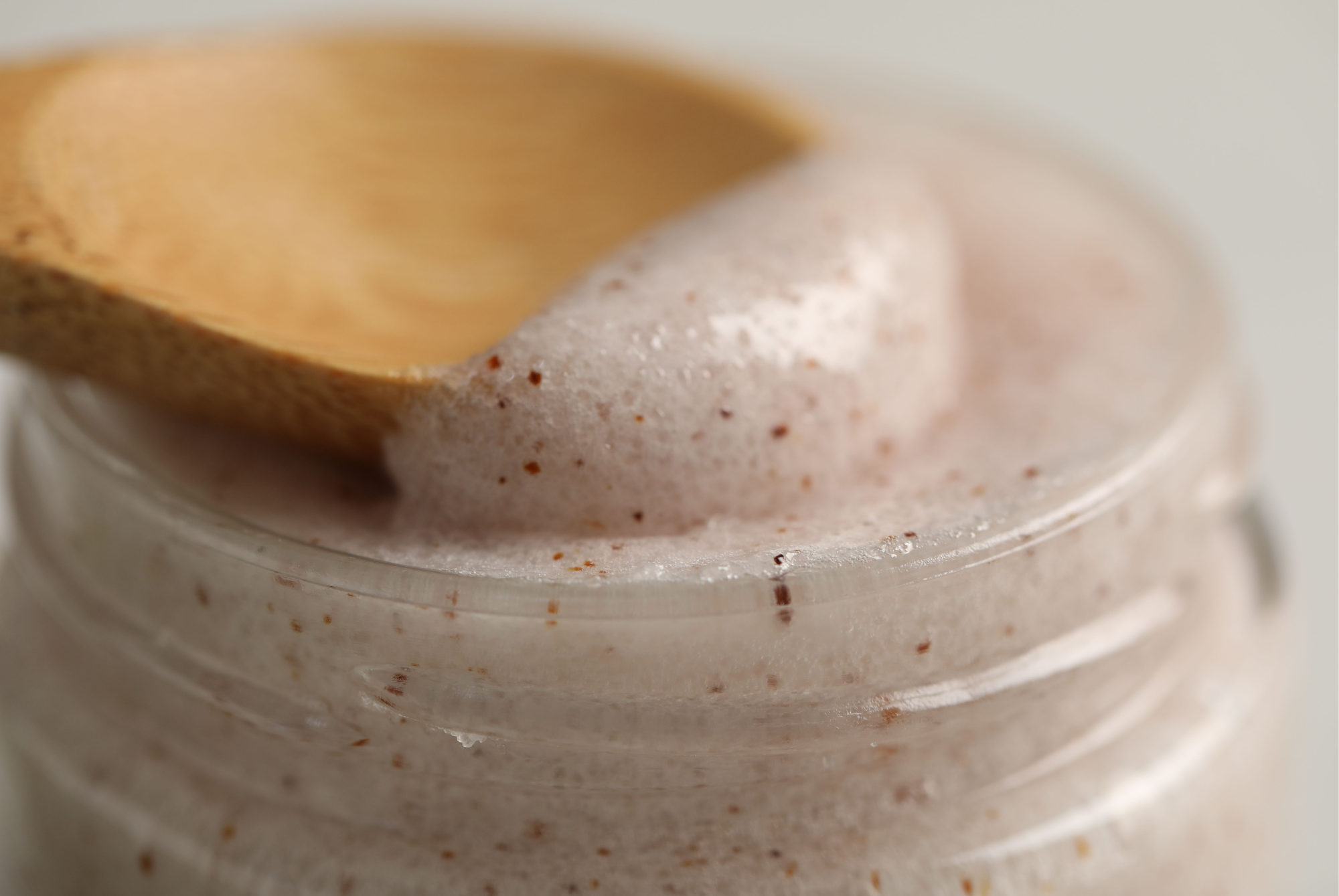A wooden spoon lifting a pale pink grainy face scrub out of an open jar