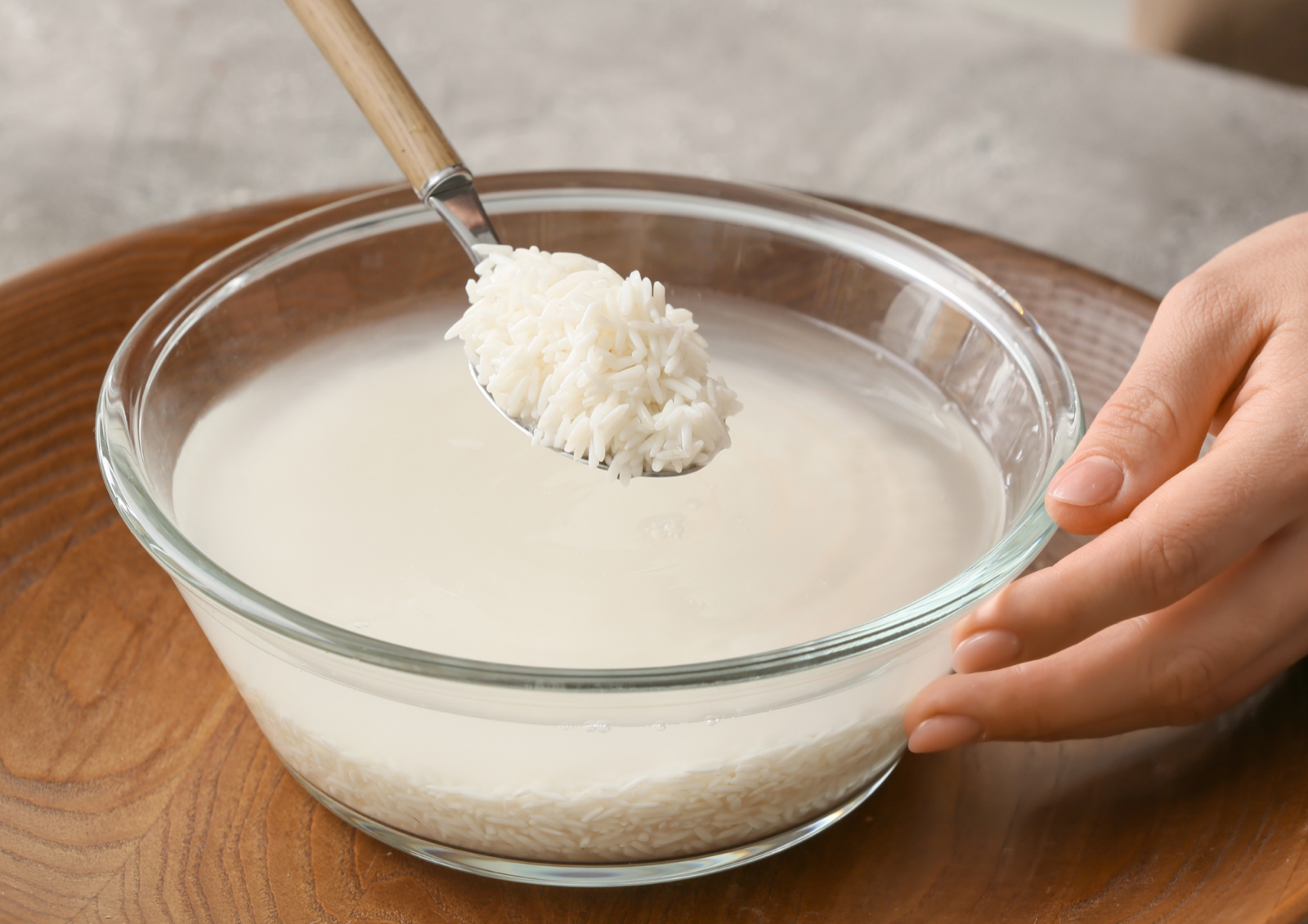 A glass bowl of cloudy rice water with a spoonful of soaked rice held above it