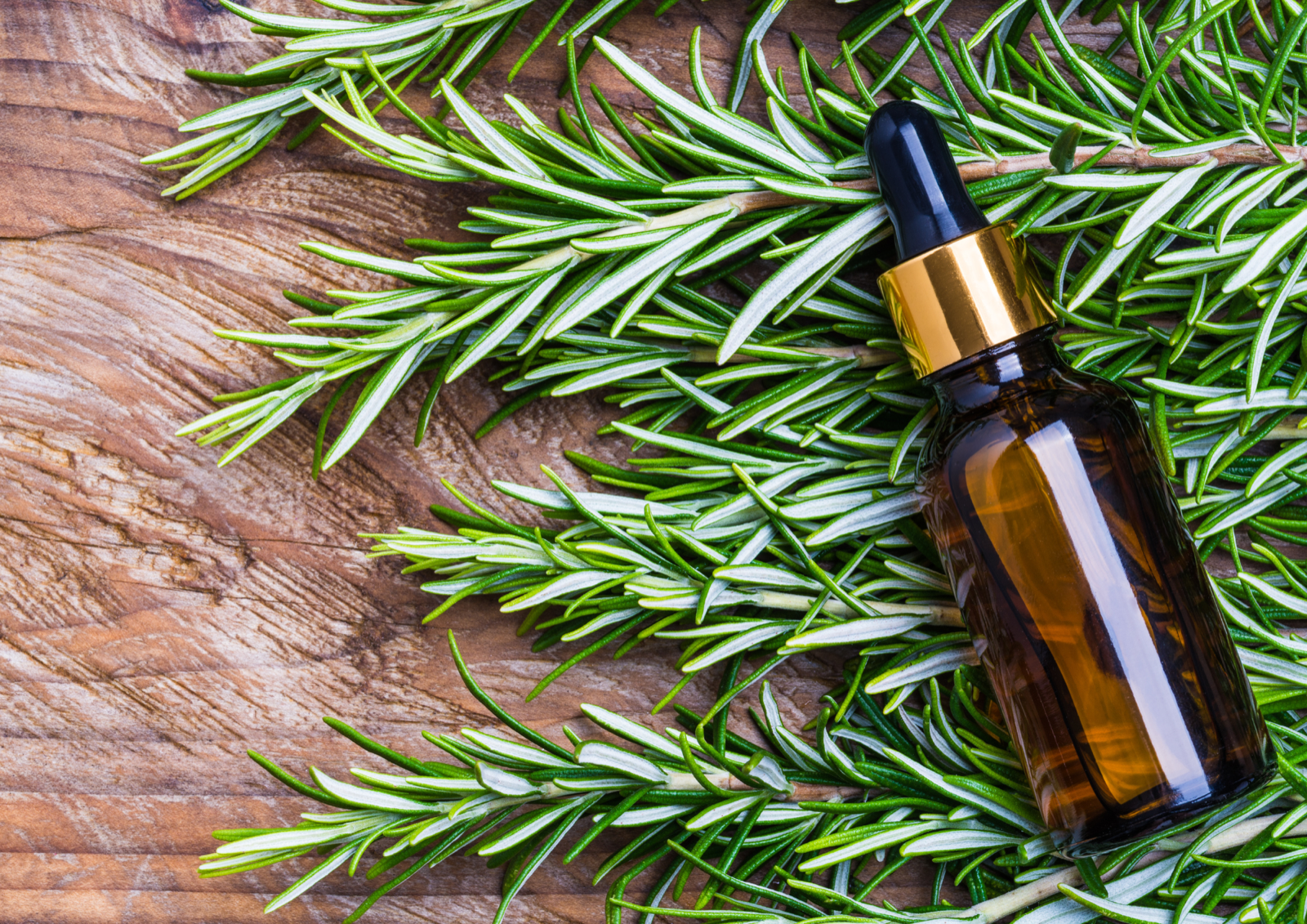 A dropper bottle of rosemary oil resting on fresh rosemary sprigs on a wooden surface