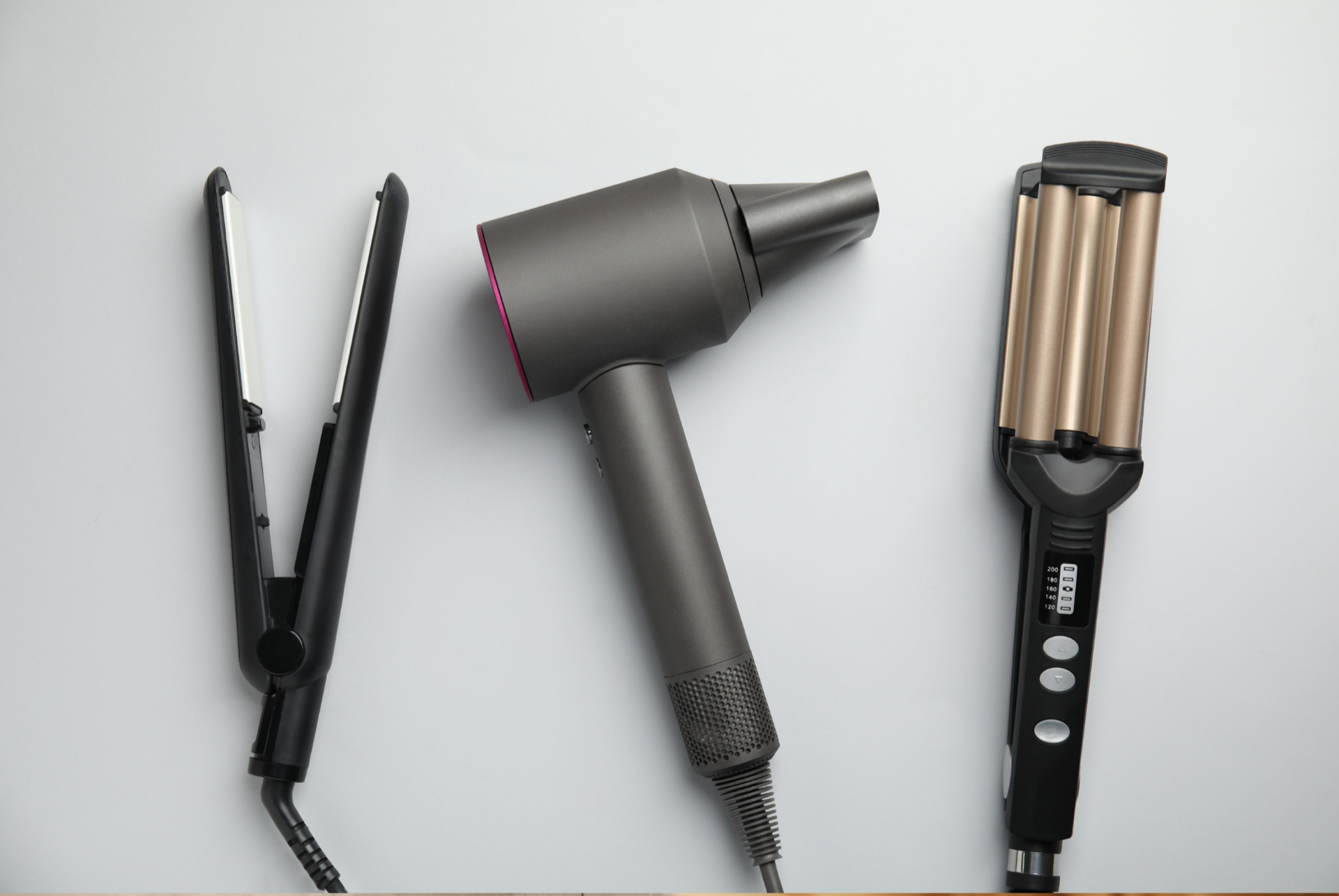 A flat iron, blow dryer, and triple-barrel waver laid out side by side on a light gray background