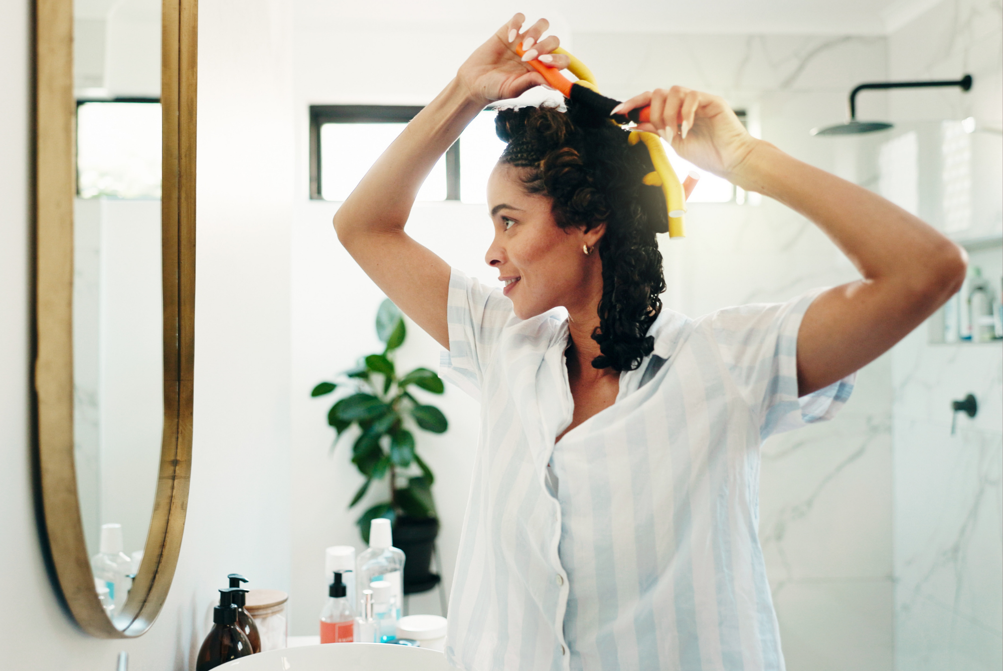 A woman in striped pajamas sectioning her curly hair with a claw clip in front of a bathroom mirror