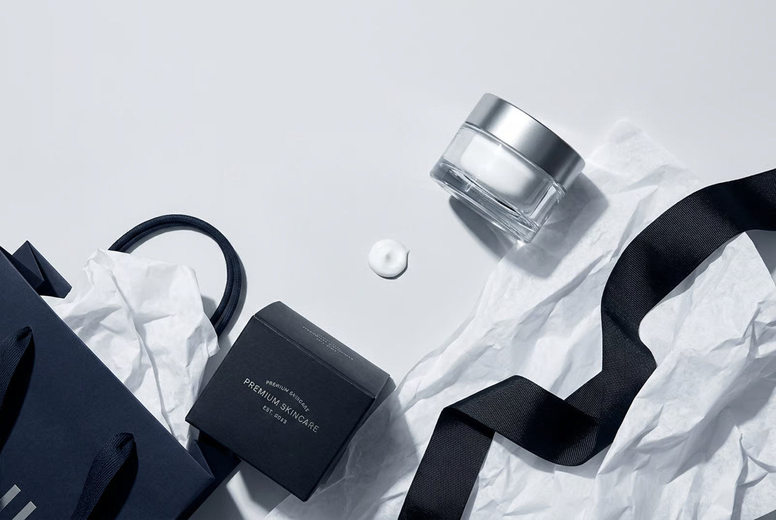 A luxury cream jar beside its shopping bag, box, and ribbon on a pale background