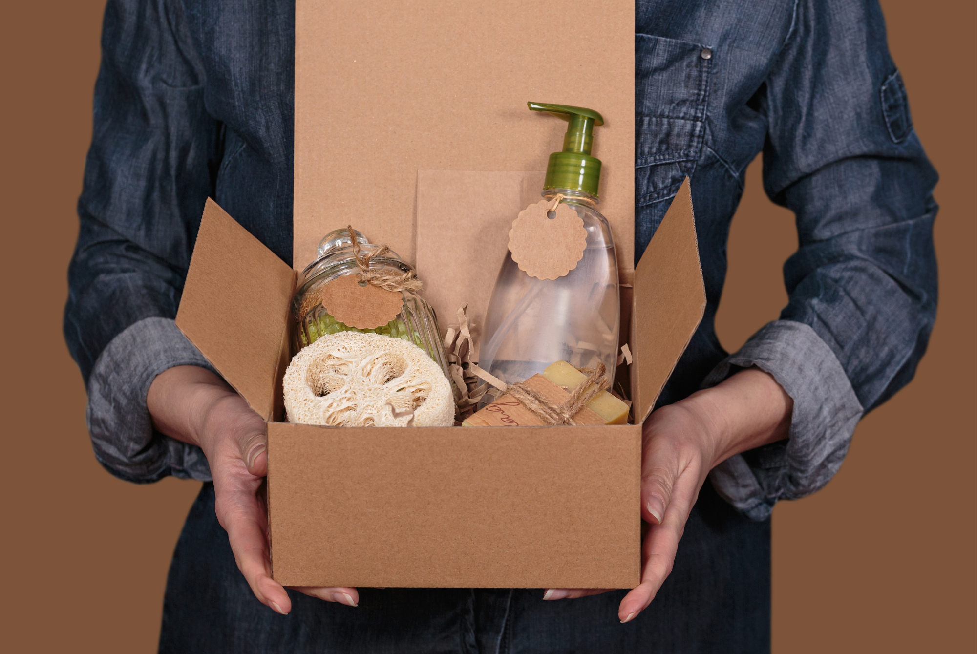 A person holding a cardboard box packed with a refill pump bottle, a glass jar and loofah sponges