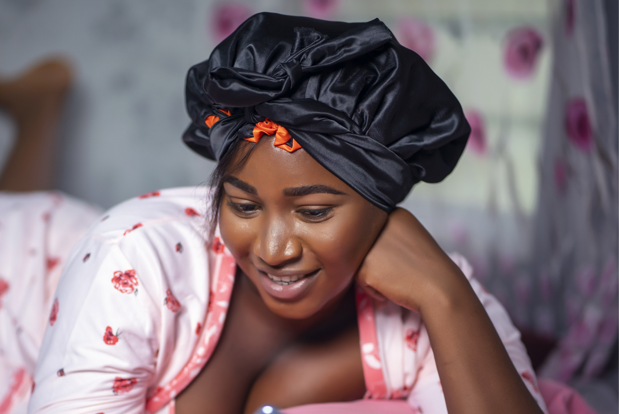 A person in pajamas lying on a bed wearing a loose black satin bonnet tied at the front