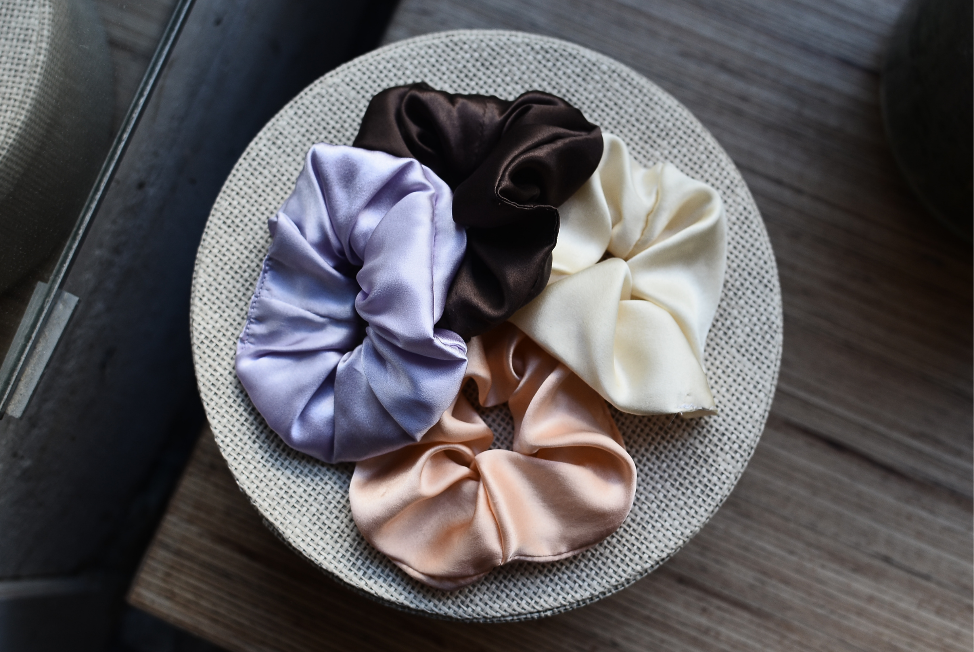 Five satin scrunchies in lilac, brown, cream, and peach arranged on a woven dish